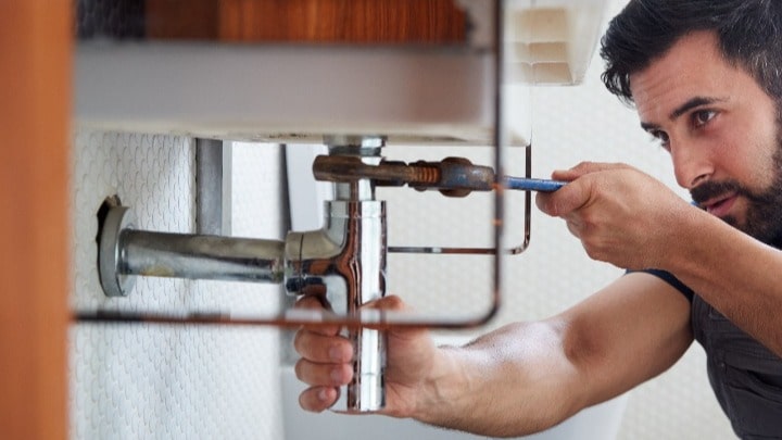 Man with beard fixing uses a wrench to fix a pipe under a sink
