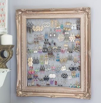 Large picture frame with chicken wire behind it with many pairs of eearings.